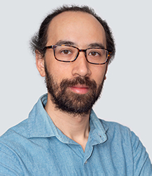 A man with dark hair, glasses, and a beard wearing a light blue button-down shirt against a gray background.