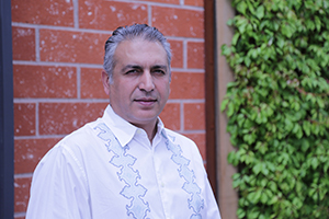 A man with graying hair wearing a white button-down shirt with light blue embroidery stands before a brick wall.