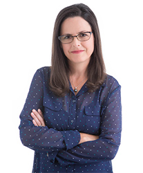 A woman with long dark hair and glasses wearing a blue polka dot blouse, posing with arms crossed.