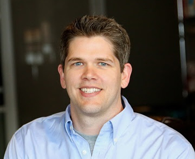 A smiling man with short brown hair wearing a light blue button-down shirt against a dark background.
