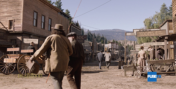 People walk along a dirt street lined with wooden buildings in an Old West town.