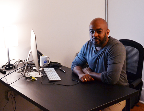 A man in a grey long-sleeve shirt sits at a black desk with a computer monitor, keyboard, and white mug.
