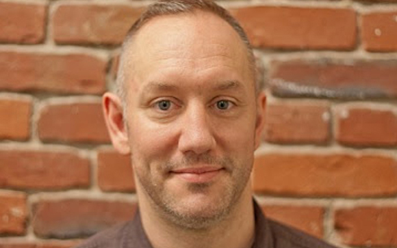 A man with light skin and short hair smiling against a red brick wall background.