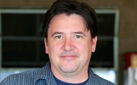 A man with dark hair and light skin wearing a blue striped button-down shirt, looking at the camera.