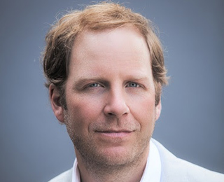 A man with wavy light brown hair and blue eyes looking directly at the camera against a gray background.