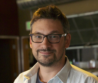 A man with short brown hair, glasses, and a light beard smiling at the camera.