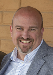 A smiling man with a bald head and a grey goatee wearing a blue blazer and checkered shirt.