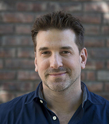 A man with short brown hair and light stubble smiling in a blue button-down shirt.