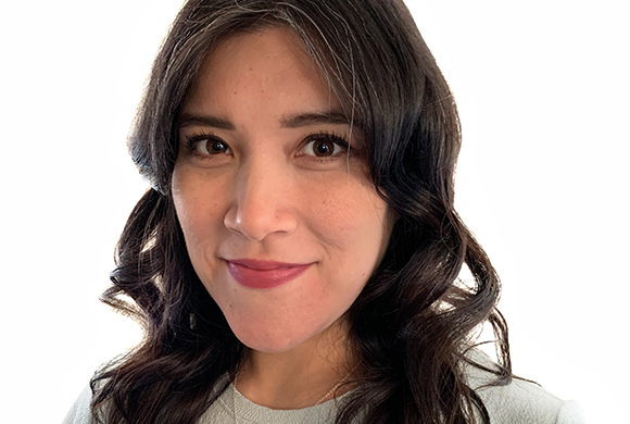 A smiling woman with dark, wavy hair and a light-colored sweater against a white background.