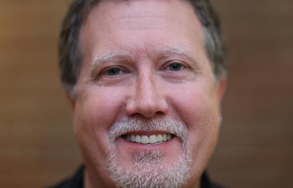 A close-up portrait of a man with light eyes and a grey goatee smiling at the camera.