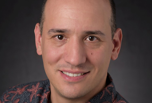 A smiling man with short dark hair and brown eyes wearing a dark patterned shirt against a dark background.