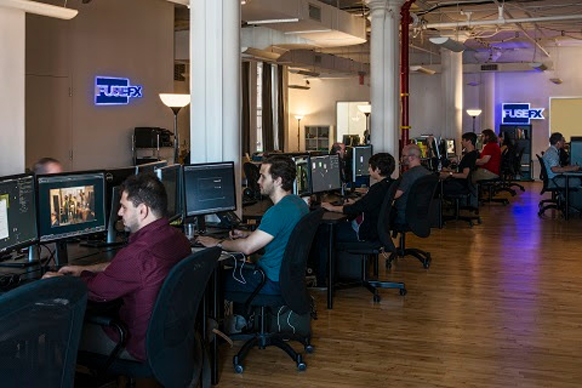 Several people sit at desks with computer monitors in a large, open-plan office with wooden floors.