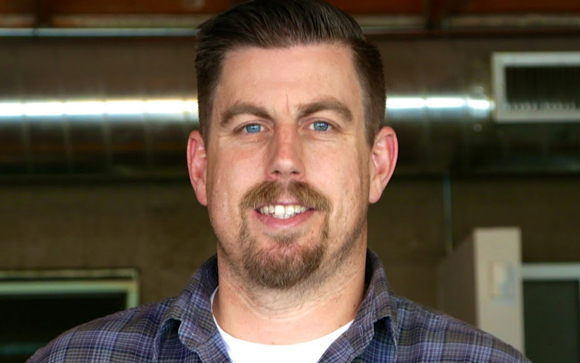 A man with short brown hair, blue eyes, and a goatee wearing a plaid shirt.