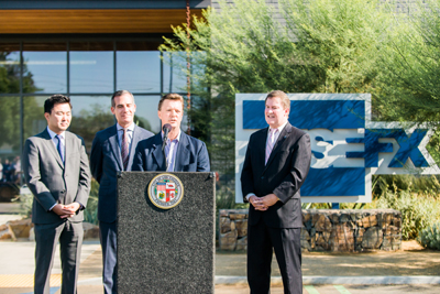Four men in business suits stand behind a black podium in front of a large blue and white sign and greenery.