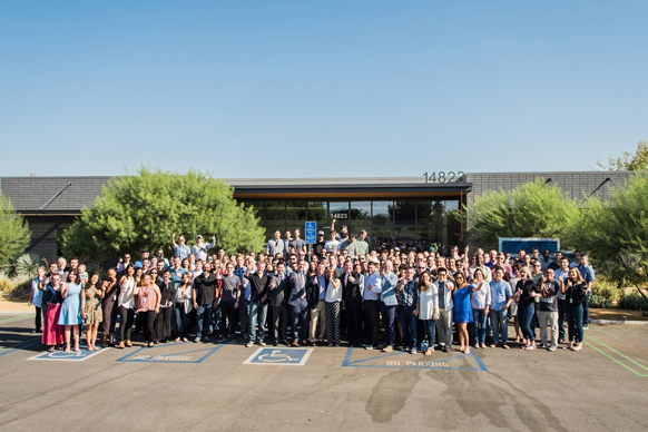 A large group of people standing together in a parking lot in front of a single-story building.