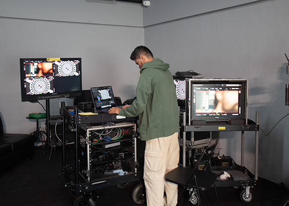A person in a green hoodie operates a laptop and monitors on mobile equipment carts in a studio.