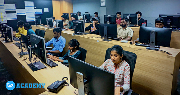 Several students sit at long desks using desktop computers in a classroom setting.