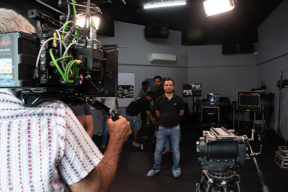 A camera operator films a man standing in a dark studio with other crew members in the background.