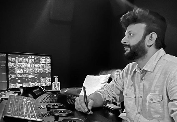 A man with a beard sits at a desk with multiple monitors and a control panel in a dark room.