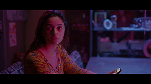 A young woman with long dark hair looks thoughtfully to the side in a dimly lit room.