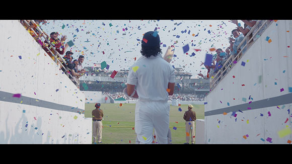 A cricket player in white stands on a field as colorful confetti falls and crowds cheer from the stands.