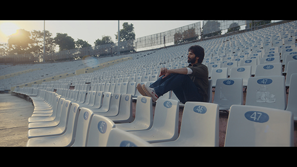 A man sits alone in rows of empty stadium seats during sunset.