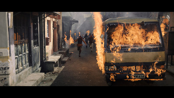 A yellow bus engulfed in flames on a narrow street with people running in the background.
