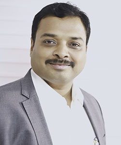 A man with dark hair and a mustache wearing a grey blazer and white collared shirt.