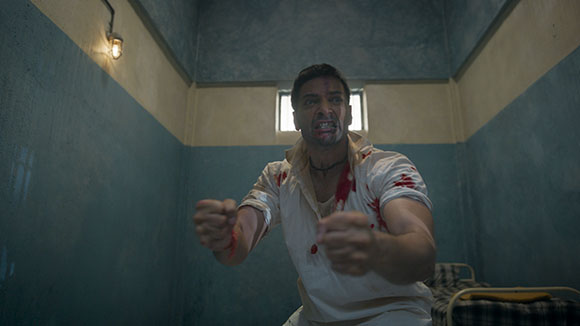 A blood-stained man in a white shirt shouts with clenched fists inside a small, dim room.