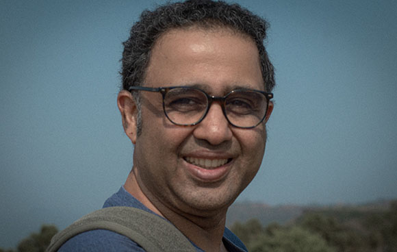 A smiling man with dark curly hair and glasses looking toward the camera against a clear sky.