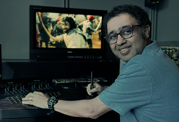 A man with glasses smiles while using a stylus and keyboard at a video editing desk with a monitor.