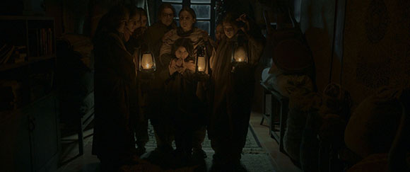 A group of people in dark clothing stand in a dim room, holding lit lanterns that illuminate their faces.