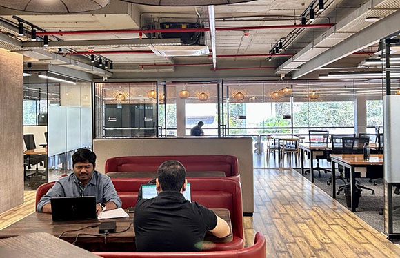 Two men sit at a wooden table with laptops in a modern open-plan office with glass partitions.