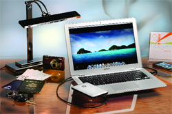 A silver laptop displaying a tropical beach scene next to a desk lamp, camera, and smartphone on a wooden surface.