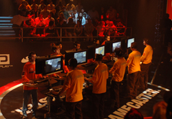 Gamers in yellow and red shirts compete at computer stations during a tournament under red lighting.