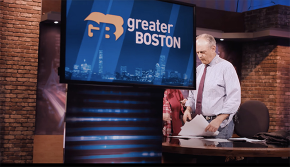 A man in a light shirt and tie looks down at papers in a studio next to a large screen with a city skyline.