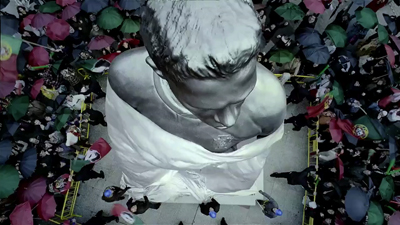 High-angle shot of a large grey statue draped in white cloth, surrounded by a crowd with colorful umbrellas.