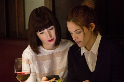 Two women sit together at a table, looking down intently at a smartphone held in one of their hands.