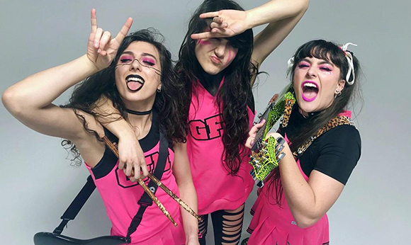 Three women in bright pink clothing posing with a guitar and drumsticks against a grey background.