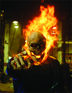 A figure with a flaming skull head and skeletal hands pointing forward in a dark setting.