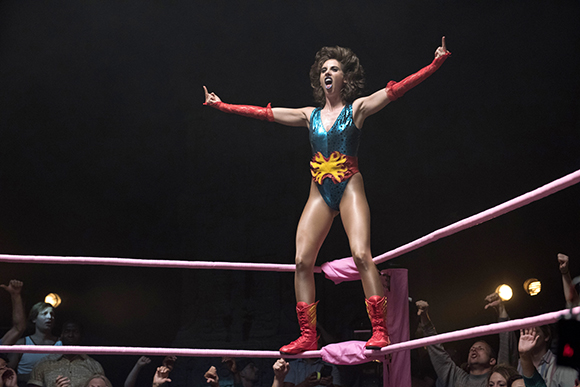 A woman in a blue sequined leotard and red gloves stands in a wrestling ring with arms raised.