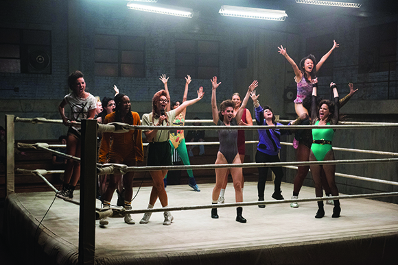 A group of women in colorful athletic wear dancing and posing inside a wrestling ring under bright lights.