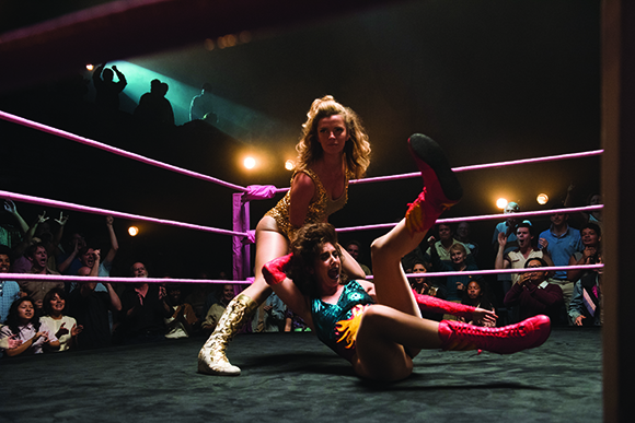 A female wrestler in a gold outfit falls backward onto the mat during a match in a dimly lit ring.