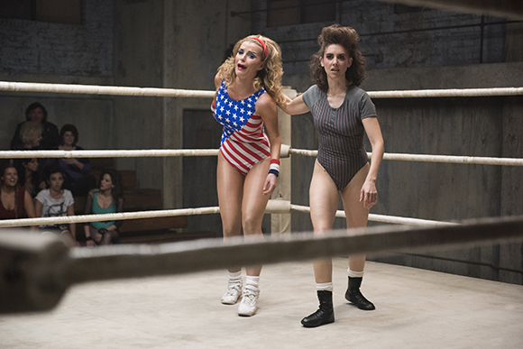 Two women in leotards stand inside a boxing ring with ropes in the foreground.