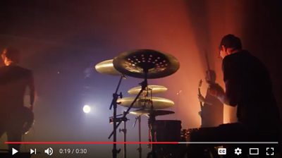 A drummer plays a drum kit on a stage under red and orange stage lighting.