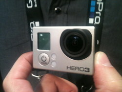 Silver GoPro HERO3 camera with lens and screen visible, held by hands. Lanyard with 'GoPro' logo visible around neck.