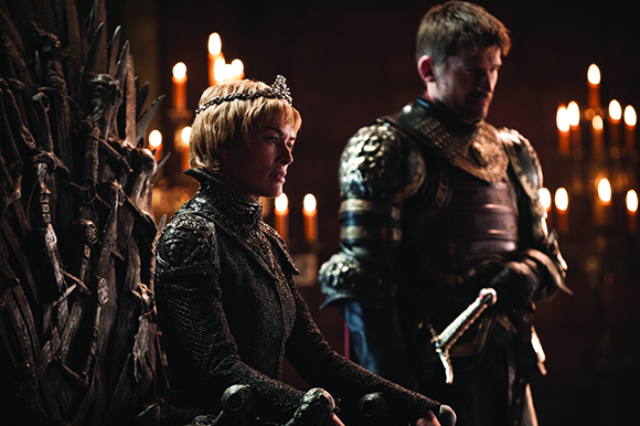 A blonde woman in a crown sits on a dark throne of swords as a man in armor stands behind her.