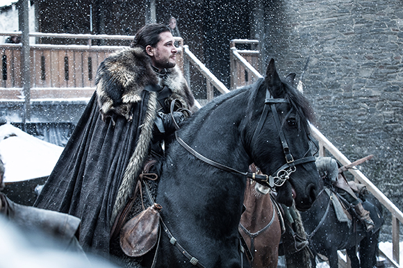 A man with dark hair wearing a heavy fur cloak rides a black horse through falling snow.