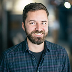 A smiling man with short brown hair and a beard wearing a plaid shirt.