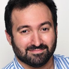 A man with dark hair and a beard smiling slightly, wearing a blue and white striped shirt.